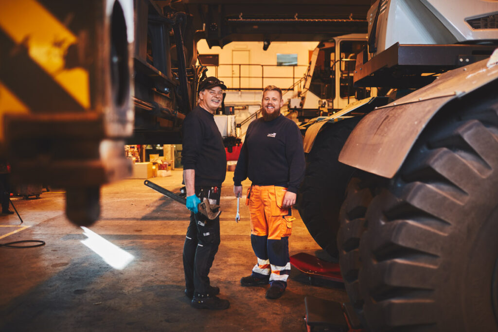 Två medarbetare från verkstaden står mitt emot varandra och ler mot betraktaren