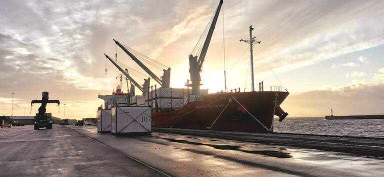 Fartyget Asian Bulker lossar husmoduler i Helsingborgs Hamn. Soluppgång bakom fartyg med kranar och husmoduler.