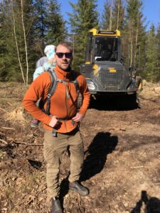 Man med barn i sele på ryggen ute i skogsmark. Skogsmaskin, träd och mark.