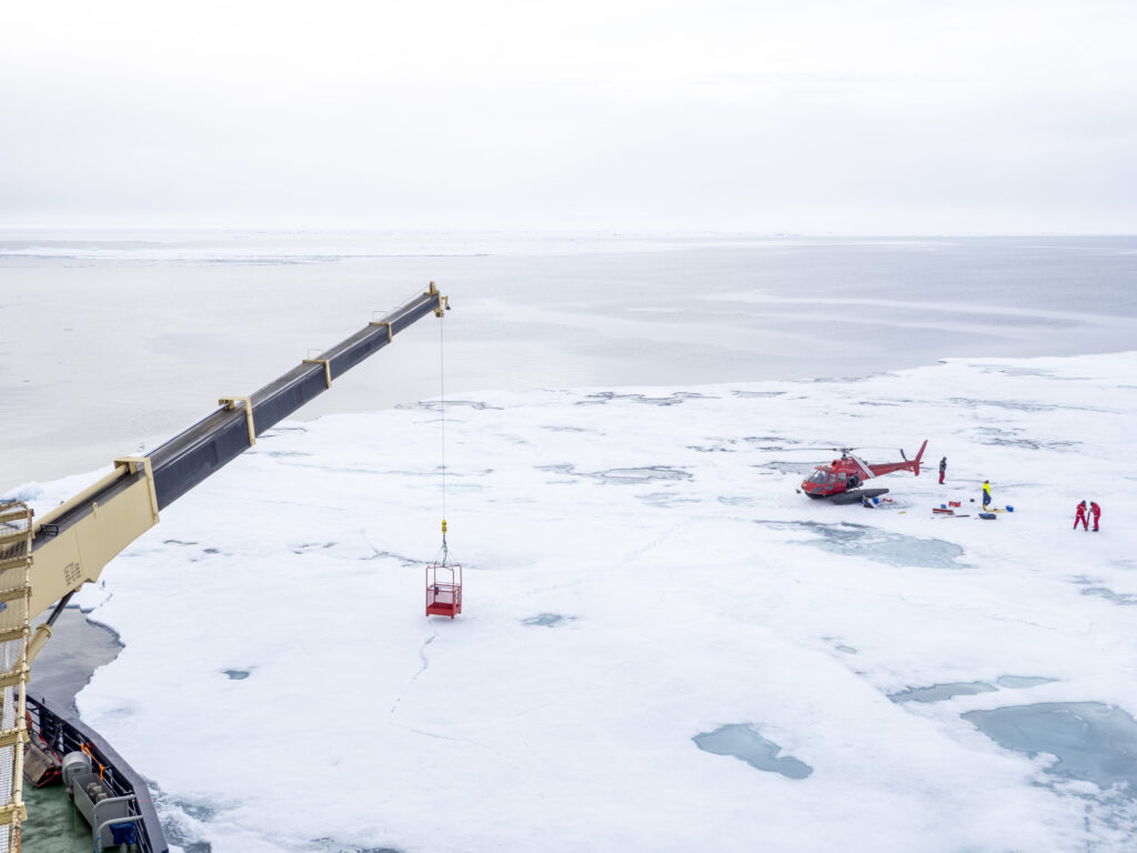 Röd helikopter har landat på isen och två personer lastar av utrustning. Vinsch med röd korg från isbrytarfartyg står på isen.