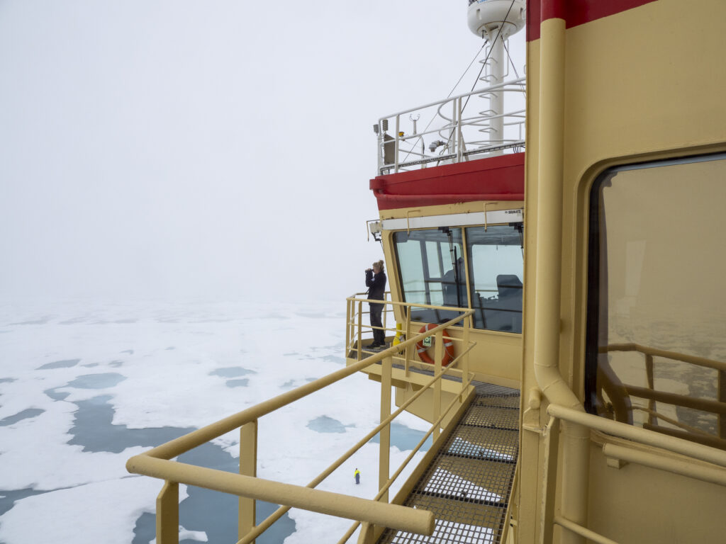 Gångbro utanför fartygsbrygga på isbrytaren Oden. Manskap står med kikare och tittar ut över isen.