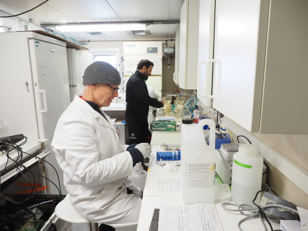 Forskare i vit rock utför tester i laboratorium. Man i svarta kläder arbetar i bakgrunden.