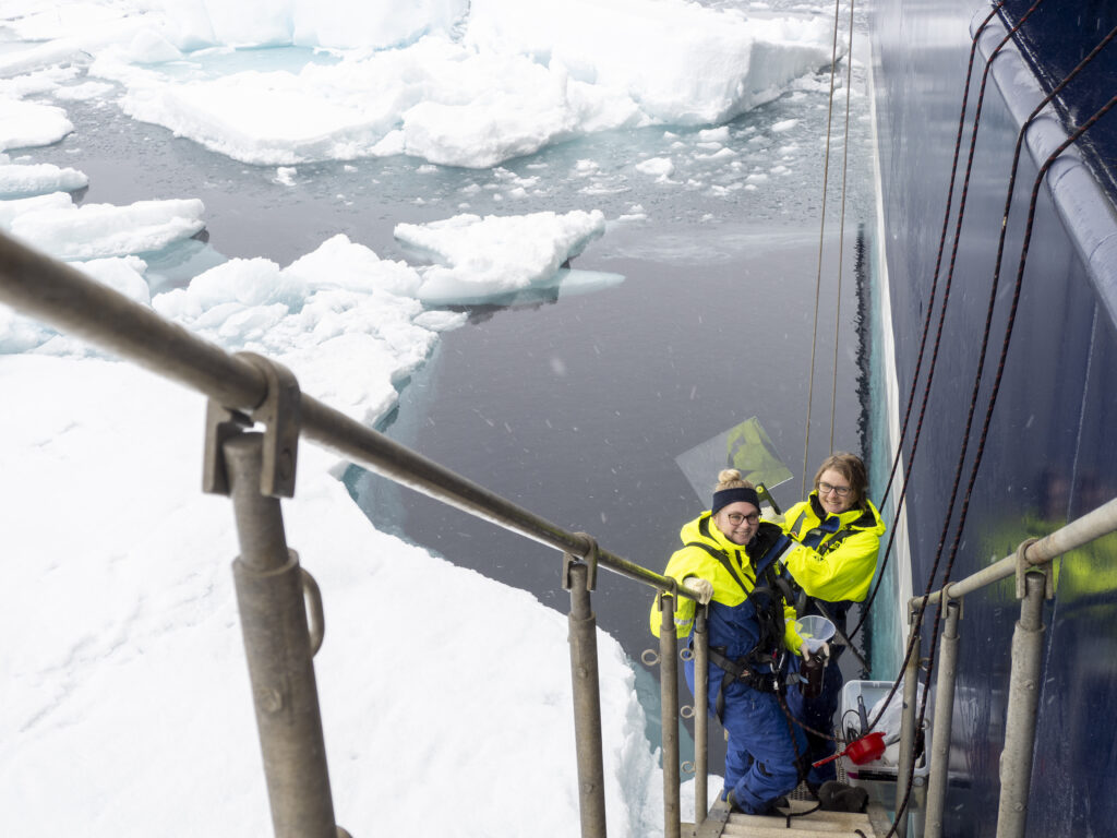 Två personer i varselkläder står i en fartygstrappa och tittar upp. Under trappan är is och hav.