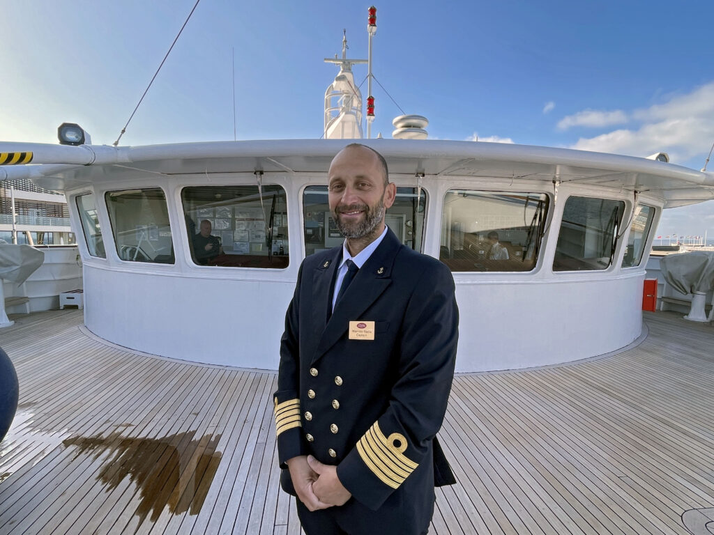 man iklädd sjökaptensuniform står på ett tik däck med båtens kommandobrygga bakom sig