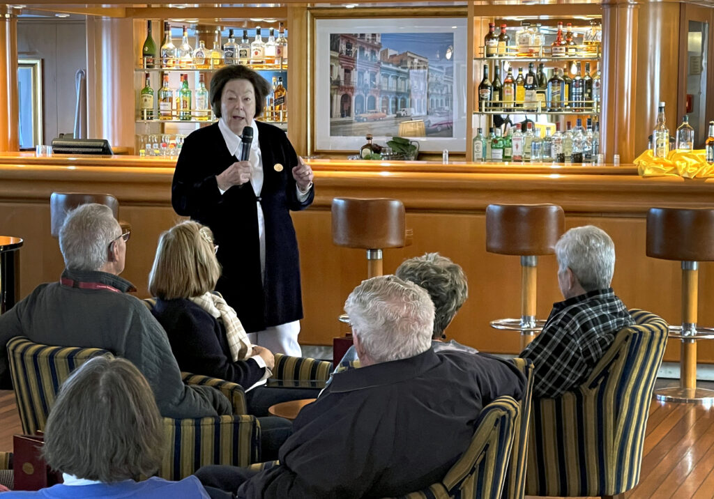 Kvinna hpller microfon och talar till publik som sitter i fotöljer framför henne. I bakgrunden syns en bardisk med flaskor