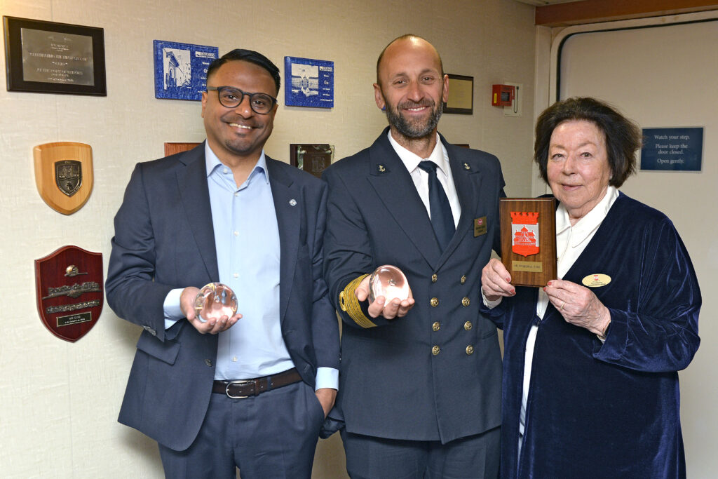 två herrar, en i mörk kostyn, en med kaptensuniform och en kvinna håller plakett och glaskulor framför en vähh med plaketter 