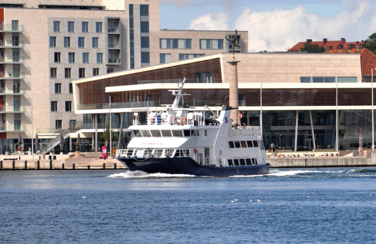 Personfärja på väg ut ur hamninlopp med stadsvy i bakgrunden.